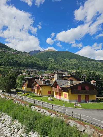 This is a photograph of a village in the mountains near Pontagna, Italy. In the foreground, a riverbed filled with large grey rocks is visible, with a dense growth of green vegetation on its embankment. A weathered wooden fence runs along the top of the embankment. Behind the fence, a paved road borders a well-maintained green lawn. Several yellow buildings with brown roofs and red shutters are situated on the lawn. These buildings have wooden balconies and stone chimneys. In the background, a verdant mountain slope is covered in a dense forest and dotted with houses. The sky above is a vibrant blue with scattered white clouds.