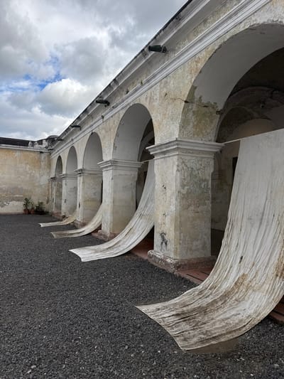 A low-angle, tilted view shows a colonnade with white stone arches supported by square columns. The stone is weathered, with peeling paint and visible cracks. Above the arches is a cornice with evenly spaced decorative fixtures. The sky with cloudy patches is visible to the left, while a gravel ground surface is seen on the right. A few small potted plants sit at the base of one of the columns.