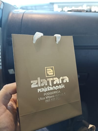 A small, golden-brown gift bag is held in someone's hand. The bag has white ribbon handles. Prominently displayed is the logo and name "zlatara majdanpek" in a stylized font, along with the address "PODGORICA, Ulica Miljana Vukova 6" and the phone number "068 888 780". The background shows the interior of a car, with the dashboard visible behind the bag.
