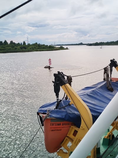 The image captures a view from a vessel on a body of water, likely a river or bay, under a cloudy sky. In the foreground, a portion of the vessel is visible, including a blue tarp covering an object and the orange side of a lifeboat or tender. The lifeboat has Cyrillic lettering that appears to read "ЕВГЕНИЙ ОРЛОВСКИЙ" and "КОТЛАС". Yellow mechanical arms, possibly davits for deploying the lifeboat, are also prominent.
The water exhibits a shimmering surface due to sunlight reflecting off the ripples. In the middle ground, a navigational buoy with red and white stripes stands in the water. The far bank of the water is lined with dense green trees and reeds. In the distance, along the far bank, there are industrial structures, including what appears to be a large crane and other buildings. The sky is filled with a mix of gray and white clouds, suggesting overcast conditions. The overall scene depicts a segment of a waterway, likely a navigable river or a coastal area, with elements of maritime equipment and infrastructure. The location is specified as Kargulino, Russia.