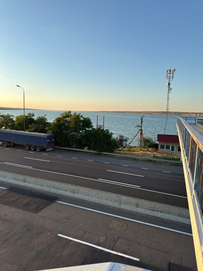 The image shows a view of a highway with a large blue truck on the right lane, heading away from the camera. The highway is next to a body of water, possibly a river or sea, with a distant shoreline visible. The sky is clear and blue, with a hint of sunset colors on the horizon. There are trees and vegetation along the embankment. A communication tower and a small building are visible on the right side of the image, near the water. A pedestrian bridge with blue railings runs along the right edge of the frame, overlooking the highway and the water. The scene appears to be located in Yelyzavetivka, Ukraine.