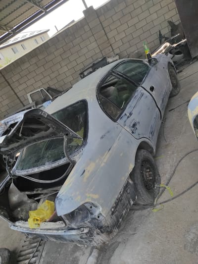 The image shows a car in a state of repair in Qairat, Kazakhstan. The car appears to be a sedan, with its bodywork covered in primer and some areas showing signs of sanding. The hood is open, revealing the engine bay, which is dusty and somewhat unkempt. A spare tire is visible next to the car, along with another detached car door, suggesting a significant amount of work is being done. The car is positioned in what looks like a garage or workshop, with concrete flooring and a rough wall in the background.
In the foreground, a green plastic bottle with a yellow cap, possibly containing some liquid, is attached to the car's side mirror. A yellow plastic bag is also visible, stuffed into a crevice of the car. The overall scene suggests a work-in-progress, with the car undergoing bodywork and possibly restoration. The lighting indicates it is daytime, but the specific time is unclear. The environment appears to be somewhat cluttered and functional, typical of a repair shop. There is no text visible in the image.