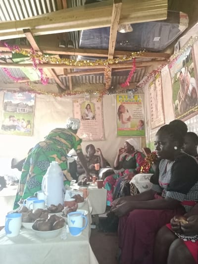 The image shows the interior of a home in Kiminini, Kenya (latitude: 0.923, longitude: 34.919). There are four women, three seated and one standing. The standing woman is wearing a green patterned dress and a white head covering. She is pouring a drink from a white thermos into a mug. There are two other mugs, one with a blue rim and one with a blue heart, on the table. There are also several small brown pastries on the table. The three seated women are dressed in various styles. One woman is wearing a white head covering and a dark red floral dress. The other two women are wearing patterned dresses. One of the seated women has a white head covering. The women appear to be drinking tea and eating pastries. The walls are decorated with various posters and pictures. The ceiling is made of corrugated metal and wood and features a blue tarp. The room is illuminated by natural light coming in from an open doorway. There are several colorful decorations on the ceiling. There is an additional, smaller, white table in the background that is out of focus.
