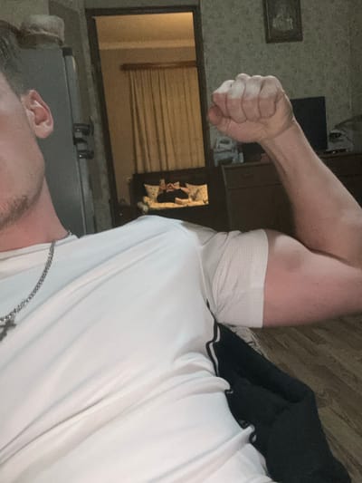 The image depicts an indoor scene within a residential setting in Durgena, Georgia. In the foreground, a male individual is partially visible, primarily his torso, right arm, and the left side of his head. He wears a white short-sleeved shirt, possibly with a mesh underarm panel, and a silver-colored chain with a cross pendant. His right arm is flexed, with the hand clenched into a fist, showcasing his bicep. The skin tone is light. The flooring beneath appears to be light wood laminate or planks.

Through a doorway in the background, another room is visible. A second individual is lying supine on a bed, which has multiple white pillows. This person is wearing dark clothing and appears to be looking down at a handheld device, possibly a mobile phone, held over their chest. Light-colored curtains are drawn behind the bed. To the right of the bed, a dark-colored dresser or cabinet supports a television, and a framed picture hangs on the wall above it. The walls in this background room exhibit a light, textured pattern.
