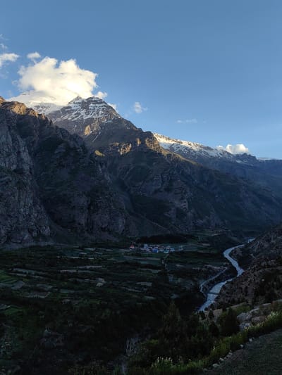 This is an aerial, eye-level, vertical shot of a majestic mountain range in Kead, India, under a clear blue sky with scattered clouds. The sun, positioned to the left, casts a warm glow on the snow-capped peaks, highlighting their rugged textures. In the foreground, a lush green valley is dotted with scattered dwellings, suggesting a small village or settlement. A winding river flows through the valley, adding a dynamic element to the serene landscape. The scene appears to be captured during the golden hour, with the sunlight creating a dramatic play of light and shadow across the mountains and the valley below, evoking a sense of tranquility and natural beauty.