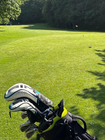 The image shows a golf bag lying on the grass of a golf course in Lancaster, UK (latitude: 54.01, longitude: -2.829). The bag is black with yellow accents and is partially open. It contains several golf clubs, two of which are in their white and black head covers. Two figures can be seen in the distance, walking on the green grass, toward the left side of the frame. The image is taken from a low angle, looking towards the bag and the golf course. The background is a field of grass, with a tree line in the background.