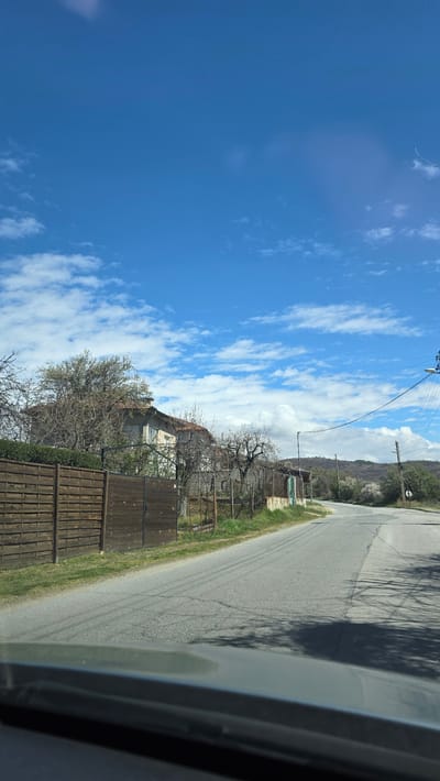 The image shows a view of a residential area, likely in Slatino, Bulgaria. A paved road runs along the right side of the frame, with a dark shadow partially obscuring its surface. A wooden fence lines the road, with trees and vegetation growing behind it. A house is visible behind the fence, and a mountain range or hill is in the distance. The sky is blue with scattered white clouds. The photograph appears to have been taken from inside a vehicle.