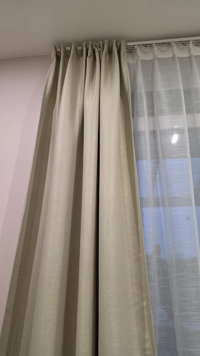 This image captures an interior view of a window, likely in a room located in Ihringshausen, Germany. The main subjects are two layers of curtains and the surrounding wall.
The window features a double-layered curtain system. The inner layer, closest to the window, is a sheer, translucent white fabric with a fine, subtle vertical striped texture. Through this sheer curtain, a soft blue sky is visible, along with indistinct, darker shapes that suggest an outdoor view of objects like buildings or trees.
The outer layer is a thicker, opaque curtain in a light beige or khaki color. This curtain is partially drawn upwards, creating prominent horizontal folds, indicative of a Roman blind style or similar gathering mechanism. It also exhibits a subtle vertical ribbed or striped texture, mirroring the pattern of the sheer curtain. Both curtain layers are attached to a white window frame or curtain rail along the top and left side of the composition.
To the immediate left of the curtains, a plain white wall is visible. Below the lower edge of the beige curtain, a wall painted in a soft, light pink hue is shown.
The lighting suggests it is daytime, with a diffused light quality implying either an overcast day or an indirect light source. There are no people, explicit activities, specific weather details beyond the sky color, or visible text present in the image.