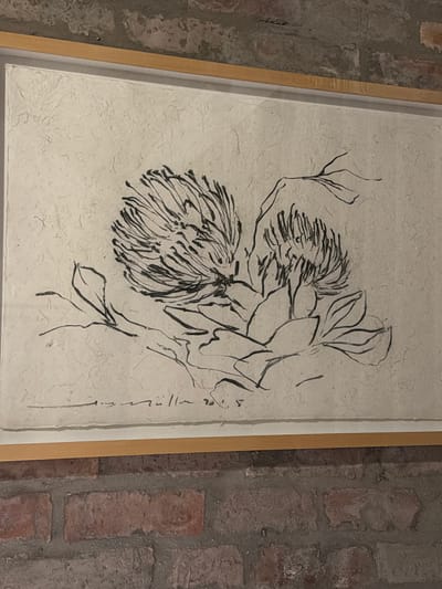A framed ink drawing on textured, light-colored paper hangs against a rustic brick wall. The artwork depicts a stylized floral arrangement, primarily featuring two large, complex flowers with numerous spiky petals or bracts, and broad leaves rendered with flowing black lines. The drawing style is organic and calligraphic, conveying a sense of movement. On the right side of the artwork, a vertical signature or inscription in script is visible, with what appears to be the number "20" at the top right. The artwork is enclosed in a simple, light-colored wooden frame with a white inner border. The background wall consists of reddish-brown bricks joined by light mortar, suggesting an indoor or sheltered setting. The lighting is relatively dim, creating a warm ambiance. The flowers bear a resemblance to proteas, aligning with the Overstrand Local Municipality, South Africa context.