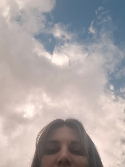 The image is taken from a low angle, looking up at a woman's face and the sky. The woman's face occupies the lower left portion of the frame. Her dark hair is visible on the left edge. The sky makes up the majority of the image, filled with white and grey clouds against a backdrop of blue. There's no information in the picture about the location being Povarenki, Russia.