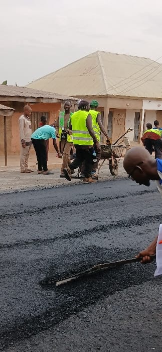 The image captures an outdoor scene depicting road construction or maintenance activities during the daytime. Several men are present, with some actively involved in the work and others observing.

In the mid-ground, a man wearing a bright neon yellow high-visibility vest and dark trousers is engaged with a metal wheelbarrow, which appears to be used for transporting materials, possibly asphalt. Another individual, partially obscured behind him, also appears to be wearing a high-visibility vest. To the left of these workers, two other men are standing and observing: one in a light blue or teal t-shirt and dark trousers, and another in a light-colored, possibly tan or beige, shirt and trousers. Further back and slightly to the right, a bald man in a blue shirt is visible. To the far right of the frame, a hand is visible holding a long, dark stick or tool, pointing towards the freshly laid dark surface.

The ground in the foreground and extending towards the right side of the image is covered with a dark, newly applied material, consistent with asphalt or tar for road surfacing. The presence of the wheelbarrow and the dark surface strongly suggests a road paving or repair project is underway.

In the background, there are several single-story or low-rise buildings with light-colored, possibly tan or brownish, walls. Some faint markings or text might be present on these buildings but are illegible. A large, light-colored roof structure, possibly corrugated metal or a similar material, with a pointed apex, dominates the upper left portion of the background, resembling a large canopy or a substantial building roof. Utility wires are visible against the overcast sky in the upper left.

The sky is bright but appears overcast, indicating daytime with diffused light. There are no prominent shadows, suggesting the sun is not directly overhead or is obscured by clouds. The overall setting appears to be an urban or semi-urban environment, likely a street or public thoroughfare undergoing infrastructural improvements.