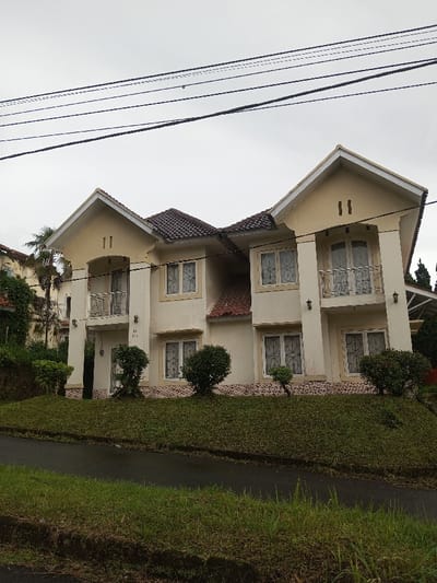A two-story duplex with a tan exterior, brown roof, and white trim is situated on a grassy hill. Balconies extend from the second story of each unit. Multiple windows with white frames are visible on both levels. Small, trimmed bushes are planted near the base of the building. Power lines stretch across the top of the frame under a cloudy sky. A paved road runs along the front of the property with grassy patches in the foreground.