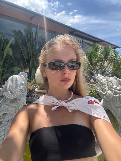 A young woman with blonde hair is taking a selfie outdoors. She is wearing a black bikini top and a white bandana with a cherry pattern tied around her neck. She is also wearing black sunglasses. The background shows a building with a corrugated roof, palm trees, and a stone statue that appears to be a dragon or mythical creature. There is a body of water in the lower right corner of the image. The sun is shining brightly, casting shadows and creating lens flare. The time of day appears to be midday, based on the strong sunlight. The woman's lips are painted red, and she is looking at the camera with a slight smile. The overall mood is casual and summery, suggesting a vacation or a day spent by the water. The sunglasses have "GUCCI" written on them. The setting is likely a resort or a private residence in Tuy An Nam, Vietnam, given the tropical foliage and architectural style.