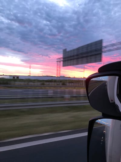 The image shows a view from a vehicle's side mirror, reflecting a sunset over a highway. The sky is filled with dramatic clouds in shades of pink, purple, and gray. In the distance, a highway stretches out, with other vehicles visible as streaks of light in the reflection. The scene appears to be on a highway near Antoninek, Poland, given the overall environment and landscape. The reflection also shows the side of the vehicle, indicating that the photograph was taken from a moving vehicle. There are no people or specific identifiable structures in the immediate foreground or reflection, but the overall context suggests a journey on a major road during twilight.