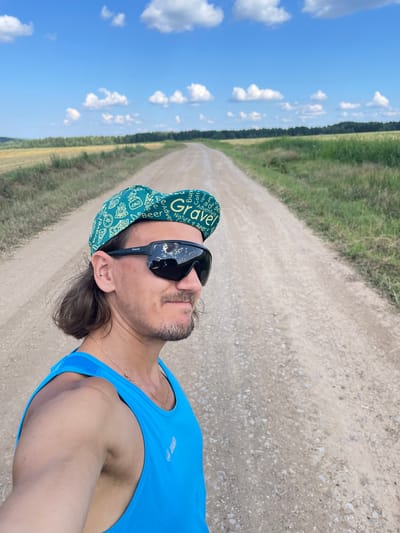 A man in a blue athletic tank top and a green cap with the word "Gravel" printed on it is taking a selfie on a dirt road. The road stretches into the distance, lined with grassy fields and trees under a blue sky with white clouds. The man is wearing black sunglasses that reflect the sky and clouds. He has medium-length brown hair and a mustache. A thin gold chain is visible around his neck. The setting appears to be rural, likely outside of Perevoznoe, Russia, given the natural landscape.