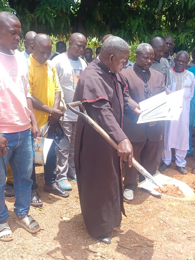 In Ninzo South, Nigeria, a group of men gathers outdoors on a sunny day. The ground is earthen. At the forefront, a man in a dark brown cassock with red trim holds a shovel filled with dirt, appearing to ceremoniously break ground. To his right, another man in clerical attire examines a stack of papers. Surrounding them are other men, some dressed casually in jeans and t-shirts, while others wear more formal attire. Their expressions are solemn and attentive. A tree with broad green leaves is visible in the background.