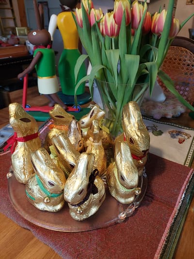 A still life features a bouquet of vibrant tulips, chocolate bunnies wrapped in gold foil, and a wooden bunny figurine. The tulips are a mix of red and yellow, with prominent green leaves. The chocolate bunnies are piled on a glass plate. The wooden bunny figurine is multicolored, with a dark brown head. The setting is a domestic interior, characterized by wooden furniture and a patterned tablecloth. The location appears to be in Ganderkesee, Germany, as the Lindt chocolate bunnies are a common Easter treat in Germany.
