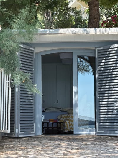 An open beach house with gray slatted shutters reveals a bedroom with a bed covered in blue and white striped sheets and a yellow and white zigzag patterned throw. A dark wood table with books sits in front of the bed. The beach house is situated on a stone-paved area with trees and foliage in the background, suggesting a location in Grad Šibenik, Croatia. The clear blue sky and reflections in the glass door indicate a sunny day.