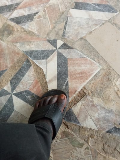 This is a close-up, high-angle shot of a person's left foot wearing a black sandal, resting on a tiled floor. The floor features a geometric pattern composed of irregularly shaped tiles in shades of beige, brown, black, and white, with some tiles appearing chipped or worn. The tiles are arranged in a mosaic-like fashion, creating a visually complex surface. The person is wearing dark-colored pants, which cover their lower leg and disappear out of frame. The lighting appears to be natural, suggesting it might be daytime, and there are no clear indicators of the weather. The scene is set in Gindin Dutse, Nigeria, suggesting an indoor or outdoor setting within that region. The focus is on the foot and the floor, with no other subjects or significant details visible. The image captures a mundane moment, with no discernible emotions expressed. There is no visible text in the image.