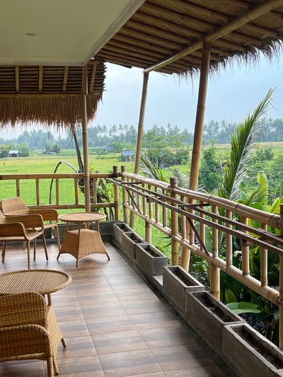 The image shows a view from a balcony in Singakerta, Indonesia. The balcony has a bamboo railing and a tiled floor with several wicker chairs and a table. The roof of the building is thatched. Beyond the balcony, there is a view of rice paddies and palm trees. The sky is overcast. There are no people visible in the image.