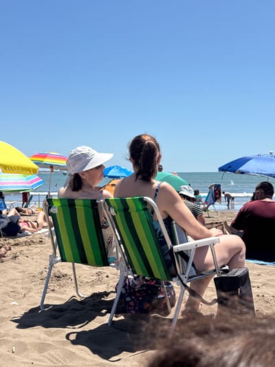 This image captures a vibrant and sunny day at the beach in Mar del Plata, Argentina, filled with people enjoying the coastal environment. The perspective is slightly elevated, looking towards the ocean.
In the immediate foreground, a woman with dark hair tied in a ponytail is seated in a green and dark-striped beach chair, facing towards the right side of the frame, her back partially to the viewer. She appears to be wearing a patterned swimsuit. Next to her, to her left, another person, likely a woman, is also seated in a beach chair, wearing a white wide-brimmed hat. Their gaze seems directed towards the sea or other activities on the beach. Strong shadows from the chairs and people are cast onto the sandy ground, indicating a bright, sunny day, likely mid-day.
Further back, towards the left, several colorful beach umbrellas are visible, including a prominent rainbow-striped one and a yellow one. Closer to the water, a large blue beach umbrella provides shade for a person wearing a maroon shirt. The ocean stretches across the midground, with gentle waves visible and several individuals in the water or near the shoreline. The sky is clear and bright blue, dominating the upper left portion of the image. The overall scene suggests a leisurely and enjoyable beach experience. There is no visible text in the image.