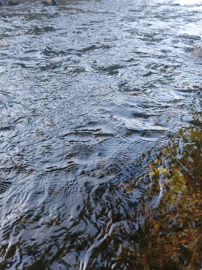 The image shows a close-up view of the flowing water of the river Emmer in Lügde, Germany. The water is clear, with ripples and currents visible on the surface. The riverbed beneath the water is composed of various sized pebbles and some aquatic plants. The lighting suggests it is daytime, with sunlight reflecting off the water. There are no people or animals visible in the frame. The focus is on the texture and movement of the water and the natural elements of the riverbed.