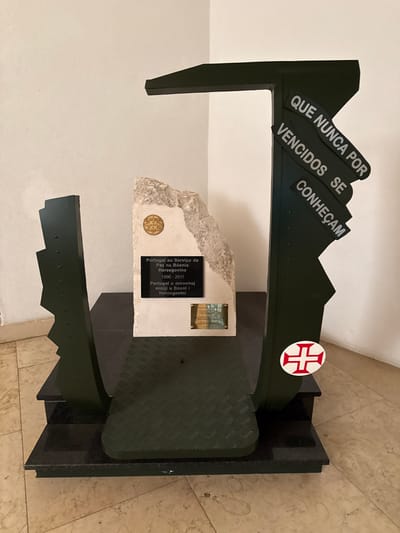 An indoor, eye-level photograph shows a commemorative monument located in the city of Praia do Ribatejo, Portugal, positioned against a plain, off-white wall in a corner. The monument rests on a light-colored, patterned tile floor.
The monument consists of a dark, multi-tiered base, topped by a dark green, rectangular platform with a diamond-plate texture. Two dark green, angular, metallic-appearing vertical structures rise from this platform. The left structure features a jagged, stepped profile. The right structure forms a prominent inverted 'J' shape with a horizontal top arm extending to the left and a vertical arm.
Centered on the platform is a light-colored, irregularly shaped stone block, suggestive of hewn rock. Embedded near the top of this stone is a circular, gold-colored medallion displaying a Portuguese coat of arms. Directly below this, a black rectangular plaque is affixed, bearing white text in both Portuguese and Bosnian: "Portugal ao Serviço da Paz na Bósnia Herzegovina 1996 - 2011" and "Portugal u mirovnoj misiji u Bosni i Hercegovini." A smaller, unreadable brass-colored rectangular plaque is positioned beneath the black one.
On the vertical arm of the right-hand dark green structure, three white horizontal strips are arranged diagonally, displaying the black Portuguese text: "QUE NUNCA POR VENCIDOS SE CONHEÇAM." Near the bottom of this same arm, a circular white emblem featuring a red Order of Christ cross is affixed. No individuals are present, and no actions or interactions are depicted.