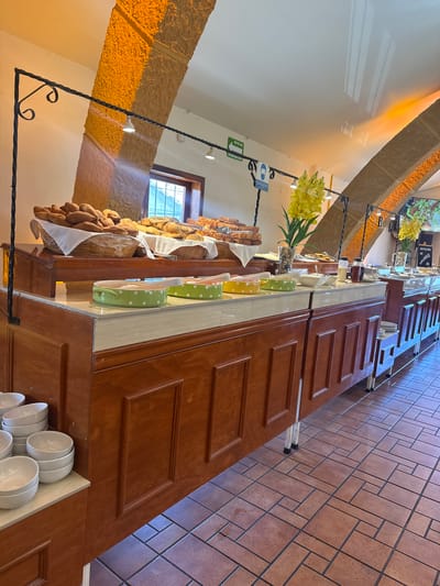 The image shows a buffet in a room with tan stone pillars and a red brick floor. The buffet table is made of dark wood with recessed paneling. Various food items, including bread, are displayed on the buffet table. There is a floral arrangement with yellow flowers on the table. The room is well-lit with natural light coming from a window and overhead lighting. White bowls are stacked at the end of the buffet. Signage is visible above the food area. Based on the architectural style and the signage, it is plausible the location is in San Cristóbal, Mexico, but there is no definitive proof from the image alone. There are no people visible in the image.