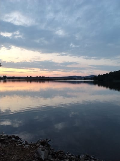 A tranquil, wide-angle view of a lake is captured at dusk. The sky is filled with a gradient of soft hues, transitioning from pale yellow at the horizon to muted blues and grays further up, interspersed with wispy clouds. The surface of the lake acts as a mirror, reflecting the sky and the distant, silhouetted treeline and rolling hills that mark the opposite shore. The foreground features a rocky, uneven embankment. The scene suggests a peaceful, natural environment, likely a rural or semi-rural setting.