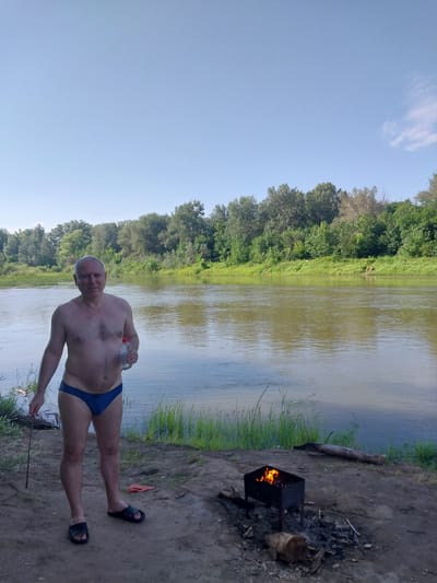 The image shows an adult male standing on the bank of a river, holding a clear plastic bottle with a white cap. He is wearing blue swimming trunks and black sandals. In the foreground, to the right of the man, is a charcoal grill with flames burning inside. There is also a log and some burnt wood on the ground near the grill. The river is wide and calm, with a tree line on the opposite bank. The sky is clear and blue. The location is the bank of the Samara River in Chkalov, Russia. The man appears to be preparing for or enjoying a barbecue by the river.