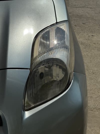A close-up, eye-level shot shows the front-right headlight of a light blue car parked on a concrete surface, possibly a street or parking lot. The car's fender curves downwards and to the right, showing a subtle reflection of sunlight. The headlight is a clear plastic casing with vertical ridges and a darker interior housing the bulb. A shadow casts diagonally across the car's hood from the left. The background consists of rough, textured concrete. The scene appears to be outdoors in daylight, possibly in Tanjungmeru, Indonesia, though no specific identifying landmarks are visible.