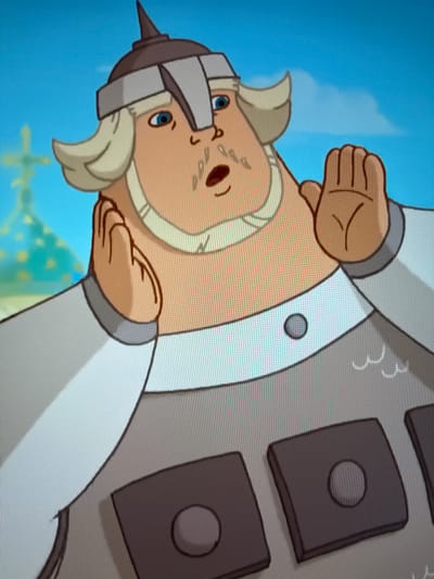 The image shows a close-up, animated, cartoon depiction of a man from the chest up, wearing a helmet and armor. The man has fair skin, light blonde hair that flares out from under his helmet, and a light blonde mustache and beard. He has blue eyes and an open mouth, as if speaking or surprised. He is wearing a dark helmet with a pointed top and a metallic band across the forehead. His armor is predominantly grey with some white accents and dark rectangular plates with circular details. His left hand is raised to his ear, and his right hand is raised in front of his face, palm facing forward. In the background, out of focus, is a building with a green dome and a cross, suggesting a church or cathedral. The sky is blue with a few soft white clouds. The style of animation and the visual elements do not correspond to any recognizable landmark or feature within the city of Oldham, United Kingdom.
