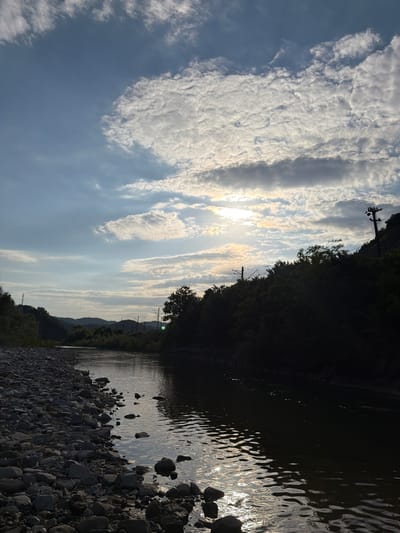 The image depicts a natural scene with a river and surrounding vegetation under a cloudy sky. The sun is visible through a break in the clouds, casting a warm glow on the water and landscape. The riverbank is lined with stones, and trees and hills form the background. There are also power lines and poles visible in the distance, suggesting human presence in the area. The scene appears to be tranquil and serene, with the natural elements dominating the composition. The location in the city of Krasnoye, Russia, is not explicitly discernible from the visual elements of the image, as it is a generalized natural landscape.