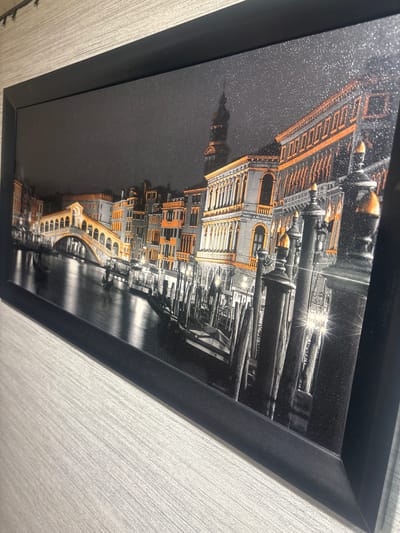 The image is a framed, black and white print with orange accents depicting a cityscape at night. The scene features a canal with several gondolas, flanked by ornate buildings and a bridge. The buildings have arched windows and balconies, with some illuminated by orange light. Lampposts line the canal, casting reflections on the water. The sky is dark and speckled, suggesting stars or a textured print. The print is framed in a black, matte finish frame. The background wall appears to be textured, possibly wallpaper, in a light grey or beige color. The image is angled, with the bottom left corner closer to the viewer. There are no people visible in the image. The location is not Маяк, Russia, but appears to be Venice, Italy, likely showing the Rialto Bridge or a similar canal scene.