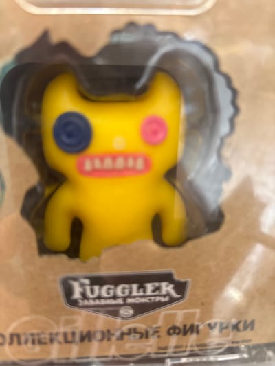 The image shows a yellow Fuggler toy with mismatched button eyes (one blue, one pink) and a jagged pink mouth with white teeth, contained within clear plastic packaging. The packaging is against a light brown, textured surface that appears to be cardboard. Text on the packaging includes "FUGGLER" in a stylized font, and below that, in smaller text, "ЗАБАВНЫЕ МОНСТРЫ" (which translates to "FUNNY MONSTERS"). Below this, larger text reads "ОЛЛЕКЦИОННЫЕ ФИГУРКИ" (which translates to "COLLECTIBLE FIGURES"). There are no people or actions depicted. The image does not provide any information about the location in the city of Kuzmolovsky, Russia.