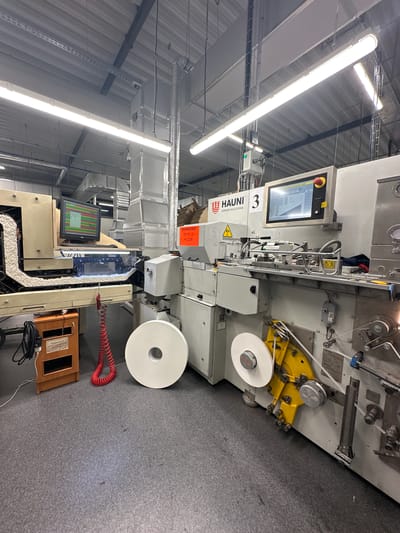 The image shows a section of a manufacturing facility in Topoli, Bulgaria. The primary focus is on two industrial machines used in the production of cigarette filters. One machine, branded "HAUNI KÖRBER SOLUTIONS 3", is light gray and features a control panel with a display screen showing data. It has rolls of white filter paper feeding into the system. A yellow mechanism with rollers and guides is visible on this machine. A warning sticker with Russian text is attached. The second machine, partially visible, has a monitor displaying a graph. A red coiled hose, a wooden box, and a black electrical cord with a chair are positioned near the second machine. The environment is an industrial setting with exposed ceiling structures, lighting fixtures, and ventilation ducts. The floor is covered in dark gray industrial carpeting. No people are visible in the image.