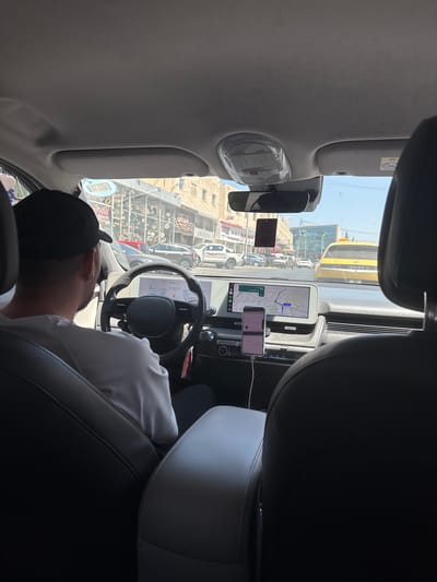A passenger's point-of-view shot taken from inside a modern car, looking towards the driver and the road ahead. The driver, a person wearing a black baseball cap and a white t-shirt, is seated behind the steering wheel of the car. The car's interior is visible, featuring a large digital display for navigation and other vehicle information, as well as a mobile phone mounted on the dashboard. Outside the car, a street scene unfolds with other vehicles, buildings, and shops, suggesting an urban environment. The yellow taxi in the background identifies the car as a taxi. The overall scene depicts a typical journey within the city of Dabouq, Jordan.