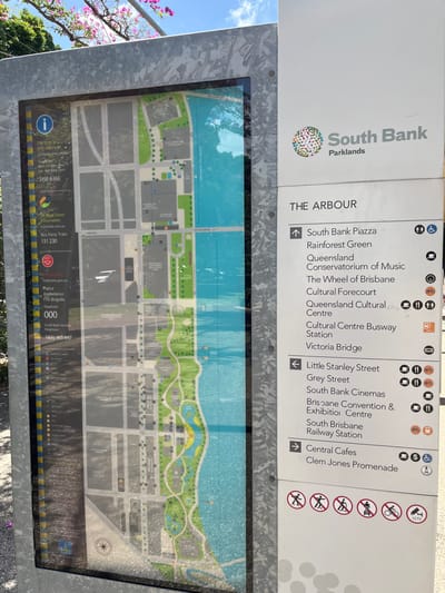 This media file captures an outdoor information sign and map for the South Bank Parklands in Brisbane, Australia, displayed on a sunny day. The sign is oriented vertically, with its top section displaying a directory and the bottom section featuring a detailed map. The sky is bright blue and clear, indicating fair weather.

The top portion of the sign features the large text "South Bank Parklands" alongside a colorful, organic-shaped logo. Below this, a directory titled "THE ARBOUR" lists various points of interest with directional arrows and small icons. Destinations pointing with an upward arrow include "South Bank Piazza," "Rainforest Green," "Queensland Conservatorium of Music," "The Wheel of Brisbane," "Cultural Forecourt," "Queensland Cultural Centre," "Cultural Centre Busway Station," and "Victoria Bridge." Destinations pointing with a left arrow include "Little Stanley Street," "Grey Street," "South Bank Cinemas," "Brisbane Convention & Exhibition Centre," and "South Brisbane Railway Station." An upward arrow also points to "Central Cafes" and "Clem Jones Promenade." Accompanying icons denote facilities like accessible restrooms, public transport, food, shopping, and no-entry signs for various activities such as cycling, skateboarding, and fishing.

The lower part of the sign is a detailed map, predominantly featuring a large blue area representing a body of water (likely the Brisbane River) bordered by green parklands and grey areas indicating roads and buildings. The map includes numerous small icons marking specific locations. In the bottom left corner of the map, there's an information icon and text for "Tourist Travel Information" (3156 6366), "Bus Ferry Train" (131 230), and "triplezero.gov.au". Emergency contact numbers for "Police," "Ambulance," and "Fire Brigade" (all 000) are also visible, along with "South Bank Security" (1300 163 847). Part of a tree with pink blossoms is visible on the far left, suggesting a lush, landscaped environment.