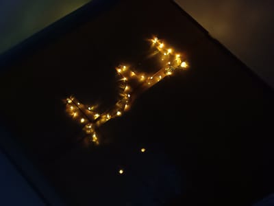 The image shows a dark room with a string of yellow fairy lights arranged in a vaguely rectangular or geometric shape on the ceiling. The ceiling appears to be composed of flat, rectangular panels. Two additional faint lights are visible in the lower center of the image. The location is unknown. There are no people present.