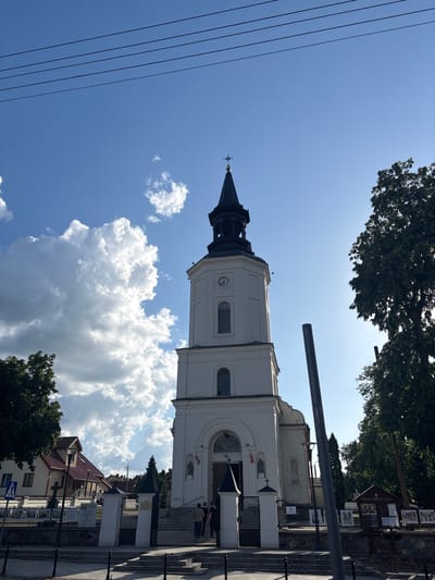 The image captures a frontal view of a white church with a tall, black-spired bell tower, situated in Sobolewo, Poland. The church's facade features a large arched entrance and a clock on the tower. In front of the church, several people are visible near the entrance. The church is framed by lush green trees on the right and left sides. In the background, a residential building with a red roof and a clear blue sky with scattered white clouds are visible. The scene appears to be a sunny day.
