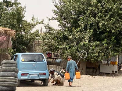 The image depicts an outdoor scene in the city of Dand, Afghanistan, characterized by unpaved, dusty ground and dense green tree foliage. In the mid-ground, a light blue Mazda Carol hatchback is parked, facing away from the viewer. A man in light-colored traditional clothing, a dark vest, and a white cap kneels beside the car's rear-left wheel, appearing to perform tire servicing. To the immediate left foreground, a stack of at least seven black car tires is visible. Behind the car and to its right, a blue horizontal air compressor tank with a red engine unit is positioned, accompanied by a circular 'no entry' sign mounted on a pole. Further to the right in the mid-ground, a second man in light blue traditional clothing walks towards the right side of the frame, carrying two orange plastic jerry cans. A third man is partially visible seated in the background to the left of the walking man. Structures include a thatched-roof shack on the far left, a brick wall behind the primary activity, and a white-fronted building or stand with yellow containers on top, located on the far right, partially obscuring a motorcycle. A small white flag or banner is discernible above the tree line in the distant background.