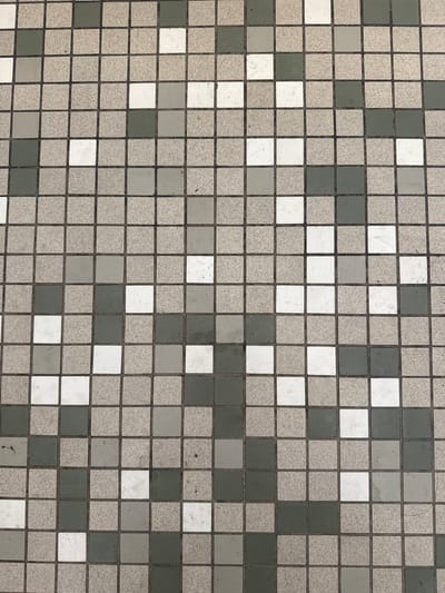 This media file is a close-up, overhead shot of a tiled surface. The tiles are small and square, arranged in a grid pattern. The color palette consists primarily of various shades of grey and white, with some tiles appearing speckled or textured, while others are smooth and solid. The grout lines between the tiles are dark and distinct, forming a clear grid. There are no people, objects, or discernible activities taking place within the frame. The lighting appears to be even, suggesting an indoor setting or an overcast day. The texture and arrangement of the tiles indicate that this could be a floor, wall, or a facade, likely in an urban or architectural context. The image lacks any text, location cues, or indicators of time of day or weather conditions beyond the ambient lighting.