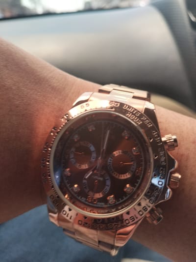 A close-up shot shows a rose gold watch on a person's wrist. The watch has a dark face with three subdials and a rose gold bezel with markings indicating "UNITS PER HOUR". The wrist and arm are visible beneath the watch. The background is out of focus, suggesting an indoor environment.