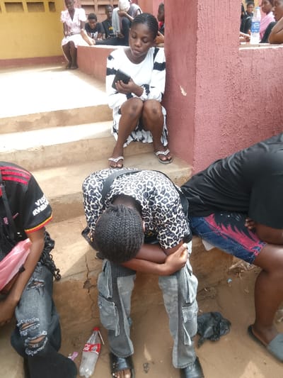 The image captures a group of individuals, primarily young adults, situated in an outdoor or semi-outdoor environment within Umuokpu, Nigeria. The setting includes concrete steps and a prominent reddish-brown textured concrete wall or pillar on the right.
In the immediate foreground, three individuals are visible:
1. To the far left, partially cropped, a person wears a black short-sleeved top with red and yellow accents, displaying "VISA RWAND" text, and ripped dark jeans. A pink plastic bag rests beside them.
2. Centrally, a person with dark braided hair, wearing a black and white leopard print short-sleeved top and grey cargo pants, is bent forward with their head resting on their arms, which are propped on their knees. An empty plastic Coca-Cola bottle with a straw lies on the ground near their feet. They wear black slide sandals.
3. To the right, a person in a black short-sleeved top and blue denim shorts with a red flame or wave pattern is partially visible, seated on the ground. A crumpled black plastic bag is near their feet, and they wear light grey slide sandals.
Slightly elevated on a concrete step, to the right of the center-foreground individual, a female subject is seated. She wears a white long-sleeved dress with broad black horizontal stripes and light-colored flip-flops with decorative straps. She is looking down intently at a black smartphone held in both hands. Her knees are visible, indicating a squatting or low sitting posture.
In the background, positioned on upper concrete steps and near the wall, several other individuals are visible, mostly seated or partially obscured. One person wears a light-colored head covering. Another individual in the background holds a clear plastic bottle of Nestle Pure Life water. A person in a light pink or white dress is also visible on an upper step to the far left of the background group. The overall scene suggests a collective gathering where individuals are resting, waiting, or quietly engaged.