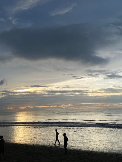 This is a vertical, eye-level, medium shot of a beach at sunset in Bateetimoh, Indonesia. The sky is filled with dramatic, dark clouds that contrast with the soft, golden light of the setting sun peeking through the horizon. The ocean is calm with gentle waves rolling onto the shore. A wide, shimmering reflection of the sunlit sky stretches across the wet sand. Two figures are silhouetted on the beach, one walking to the right and the other standing facing left, both appearing as dark shapes against the bright water. The overall mood is serene and contemplative.
