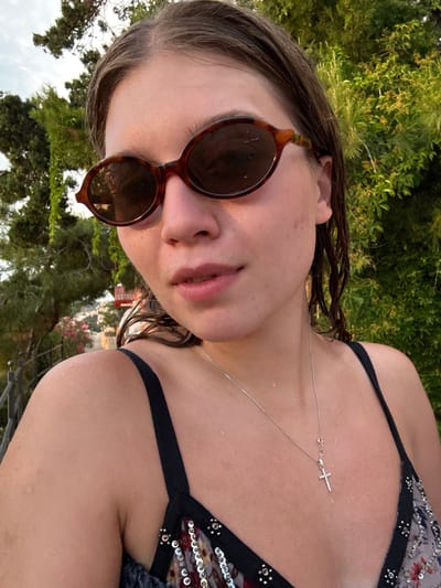 A young woman wearing sunglasses and a bikini poses for a selfie in Primošten, Croatia. The woman has wet brown hair and is wearing a silver necklace with a cross pendant. She is standing outdoors with lush green foliage and trees in the background. The sky is visible above the trees.