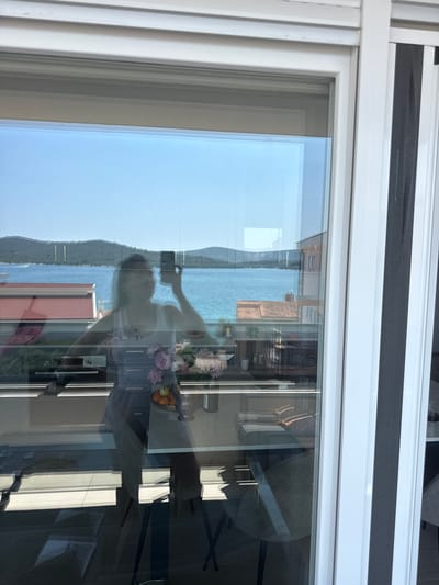 This is a photograph taken from a window, likely in Šibenik, Croatia, as suggested by the location context. The image is oriented vertically. It shows a reflection on the window, with the actual scene visible through the glass.
In the reflection, a person is seen taking a selfie with a smartphone. They are positioned in what appears to be an interior space, with a table in front of them holding a vase of flowers and a bowl of fruit. A dining table and chairs are also visible in the reflection. The interior space seems to be a well-lit room, possibly a dining area or living room, with windows and furniture.
Through the window, a scenic view of the Adriatic Sea and a coastline dotted with hills and buildings is visible. The water is a vibrant blue, and the sky is clear and bright, suggesting a sunny day. The architecture in the background, with terracotta-tiled roofs, hints at the Mediterranean style typical of the region.
The overall impression is one of a bright, pleasant day in a coastal location. The activity of taking a selfie suggests a personal moment of capturing the experience. The scene combines an indoor setting with a beautiful outdoor landscape, viewed through the reflective surface of a window.