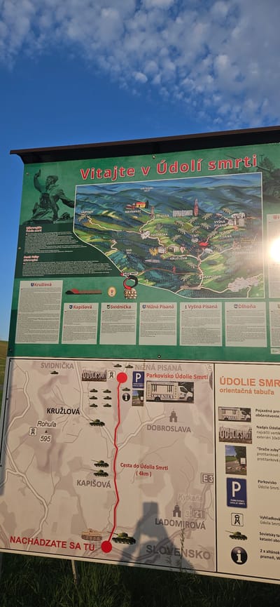The image displays an information panel about the "Valley of Death" (Údolie smrti) in Svidník, Slovakia. The panel is divided into two main sections. The upper section features a large, colorful map of the region, highlighting historical locations and showing numerous military vehicles, likely tanks, placed along a route. The title "Vitajte v Údolí smrti" (Welcome to the Valley of Death) is prominently displayed in red. Text boxes on the left and bottom of the upper section provide historical information in Slovak.

The lower section of the panel presents a more detailed map of the immediate area, with a red line indicating a path leading to "Cesta do Údolia Smrti" (Path to the Valley of Death), noting it is 4km long. This map shows village names like Svidníčka, Kružlová, Rohuľa, Nižná Písaná, Dobroslava, and Ladomirová. It also depicts several military vehicles on the map. Icons indicate a parking area ("PARKOVISKO ÚDOLIE SMRTI") at Nižná Písaná, a tourist information point, and a viewing platform. The phrase "NACHÁDZATE SA TU" (You are here) with a red marker points to a location southwest of Kružlová, near Rohuľa and a number 595.

To the right of the lower map, there is a vertical information section with titles like "ÚDOLIE SMRTI orientačná tabuľa" (Valley of Death orientation panel). It includes images of a souvenir shop, a sculpture reading "ÚDOLIE SMRTI", and a tank. Text descriptions mention "Pojazdná predajňa občerstvenie" (Mobile shop for refreshments), "Nadpis Údolie smrti najväčší vertikálny exteriéri 10x3" (The inscription Valley of Death largest vertical exterior 10x3), and "Dráče zuby" (Dragon's teeth), likely referring to anti-tank obstacles. It also mentions the parking area at Údolie Smrti and a viewing platform.

The overall scene is outdoors, with a bright blue sky and some clouds visible above the information panel. Green grass is present at the bottom of the image, suggesting a natural outdoor setting for the panel. The panel appears to be made of metal or a similar weather-resistant material and is likely mounted on posts, though only the display surface is fully visible. The overall impression is an informational exhibit detailing a historical site related to conflict and remembrance.
