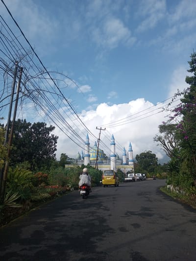 The image depicts a street scene in Mya Sit Kaing Ywar Thit, Myanmar, dominated by a large mosque with blue and yellow architectural details. A two-lane paved road curves gently into the distance. A person on a motor scooter, clad in a light-colored rain jacket and helmet, rides away from the camera on the left lane. A yellow car is also travelling in the same direction. Several parked vehicles can be seen further down the road, near the mosque. Lush greenery borders both sides of the road, including trees with purple flowers on the right. Overhead power lines crisscross the sky, partially obscuring the blue sky with scattered white clouds. A utility pole is visible on the left side of the road. The road surface appears wet, reflecting light.