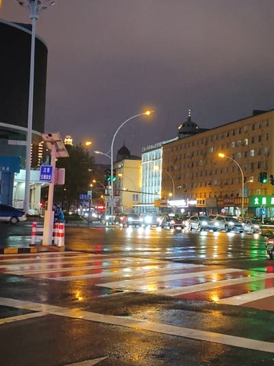 This is a photograph taken at night in Jixi, China. The scene depicts a wet street with traffic lights and vehicles. The road surface is reflective, showing the lights of cars and streetlights. In the foreground, a pedestrian crossing with white and red stripes is visible, glistening with moisture. Several cars are stopped at the intersection, their headlights illuminated. Traffic lights are showing green for vehicles heading forward.

To the left of the image, there is a tall lamp post and a sign that reads "注意 交通安全" (Zhùyì jiāotōng ānquán), which translates to "Attention: Traffic Safety." A person in a blue jacket is standing near this sign, leaning against a pole.

In the background, there are multi-story buildings. One building on the right is a yellowish-brown color with many windows. Another building on the left is lighter in color. The sky is overcast and dark, contributing to the nighttime atmosphere. The overall impression is a city intersection at night during or after rainfall, with active traffic.